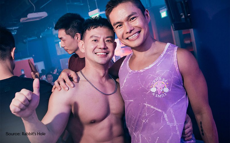 two smiling men posing together in a gay nightclub one shirtless giving a thumbs-up and the other wearing a tank top with a cartoon print