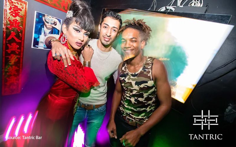 three friends posing at Tantric Bar, a drag performer in a red outfit with dramatic makeup, a man in a white shirt, and another in a camouflage tank top smiling under colorful club lights