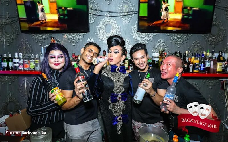 group of five people posing behind the bar at Backstage Bar, two drag performers in glamorous makeup and outfits alongside three smiling men holding alcohol bottles