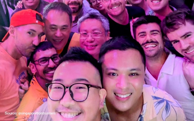 group selfie of smiling friends at a gay bar, with several men posing closely together under pink lighting