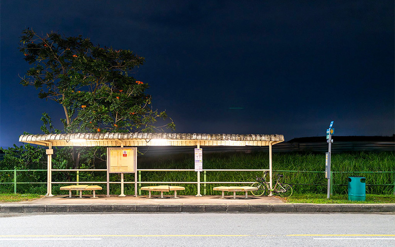 Empty rural bus stop at night with a lit shelter, wooden benches, a parked bicycle, and a large tree beside tall grass under a dark sky