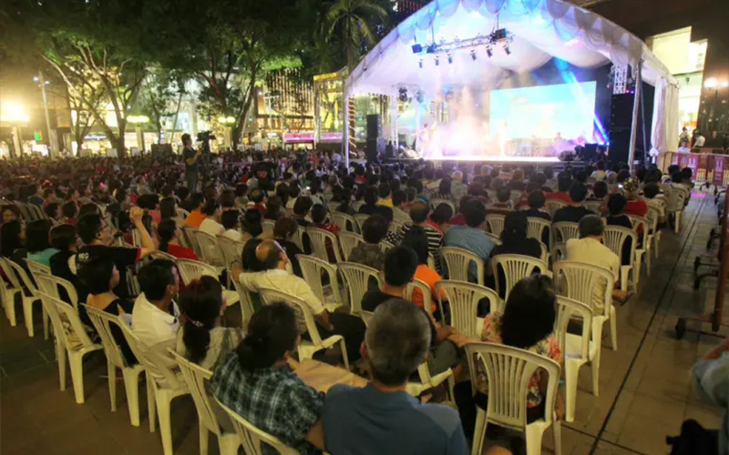 Large crowd seated on white plastic chairs watching a live stage performance under a lit outdoor tent at night