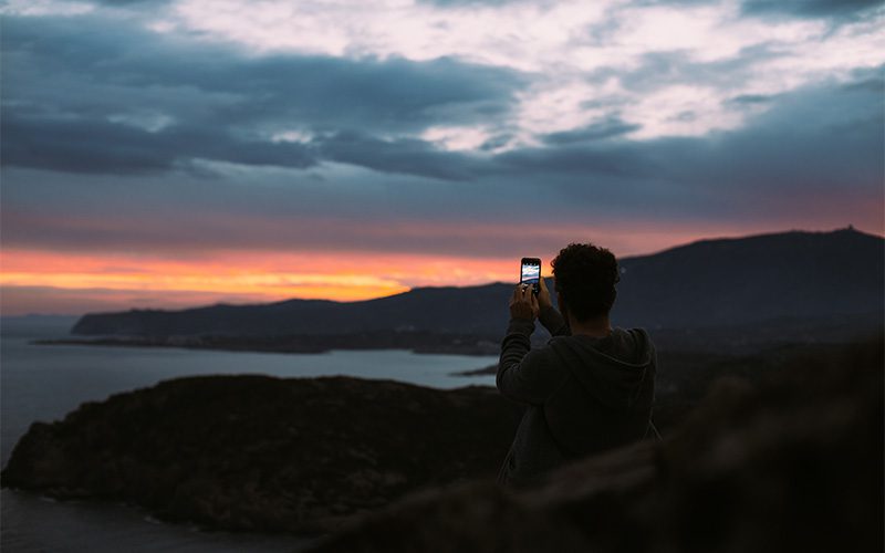 Person taking a photo with a smartphone of a colorful sunset over mountains and the sea.