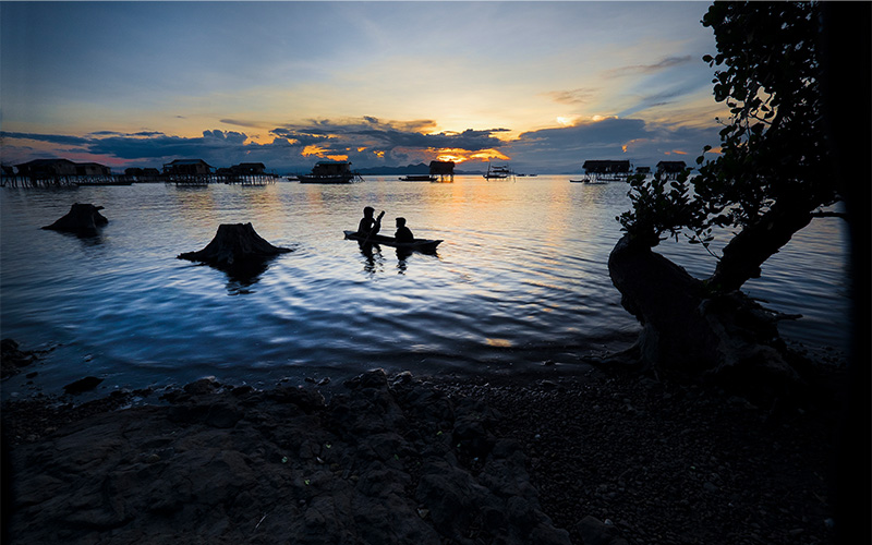 Silhouettes of two people paddling a small boat on calm water during sunset, with stilt houses and tree stumps in the background