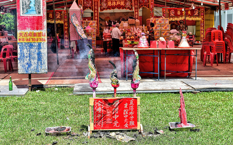 Traditional Hungry Ghost Festival altar with large incense sticks burning in front of a red tent filled with offerings, decorations, and empty red chairs